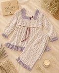 A two-piece baby pajama set featuring a long-sleeve top and matching pants in an off-white fabric with a small purple floral print, decorated with purple ruffle trim and a dark purple tie belt. The set is styled in a flat-lay arrangement on a beige fabric background with decorative props including a book, dried flowers, and a candle.
